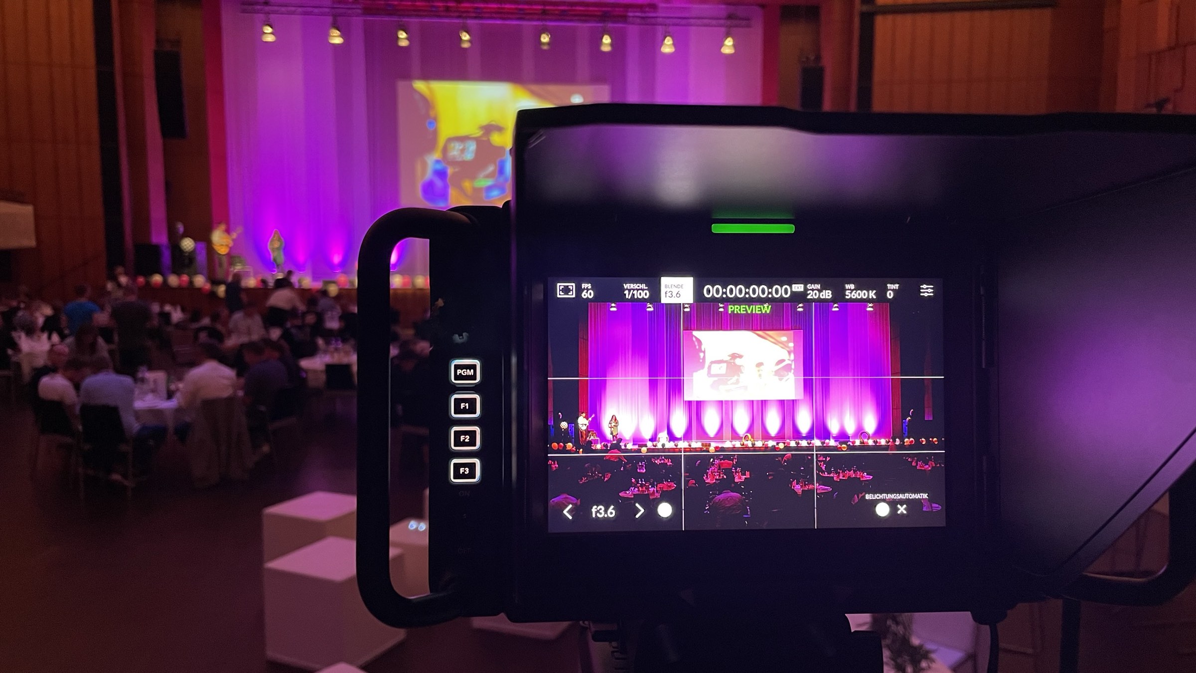 Kameramonitor mit Live-Vorschau einer Bühne in violettem Licht, im Hintergrund das Publikum eines Saals